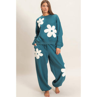 A woman modeling a teal two-piece lounge set with oversized white daisy flower appliques, consisting of a long-sleeve sweatshirt and matching drawstring sweatpants, against a plain beige studio background.