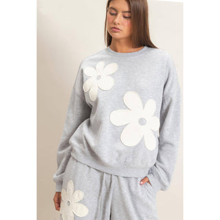 A woman wearing a light gray heather sweatshirt and matching sweatpants set, both decorated with large white fabric flower appliqués, against a plain beige studio background.