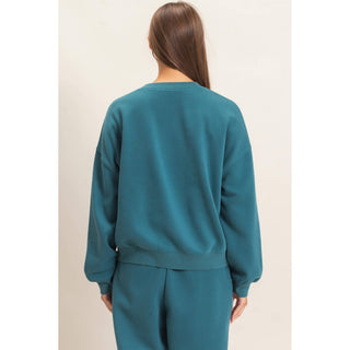 A woman viewed from the back wearing a teal crewneck sweatshirt and matching sweatpants against a plain light beige studio background.