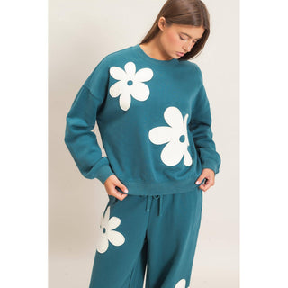 A woman wearing a teal sweatshirt and matching sweatpants set, both decorated with large white flower graphics, against a plain light beige studio background.