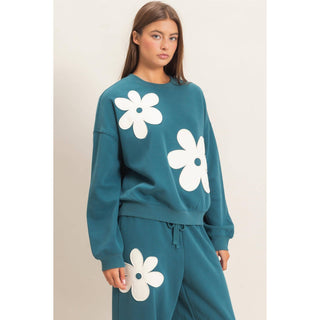A woman modeling a teal sweatshirt and matching sweatpants set, both decorated with large white flower appliques, against a plain light beige studio background.